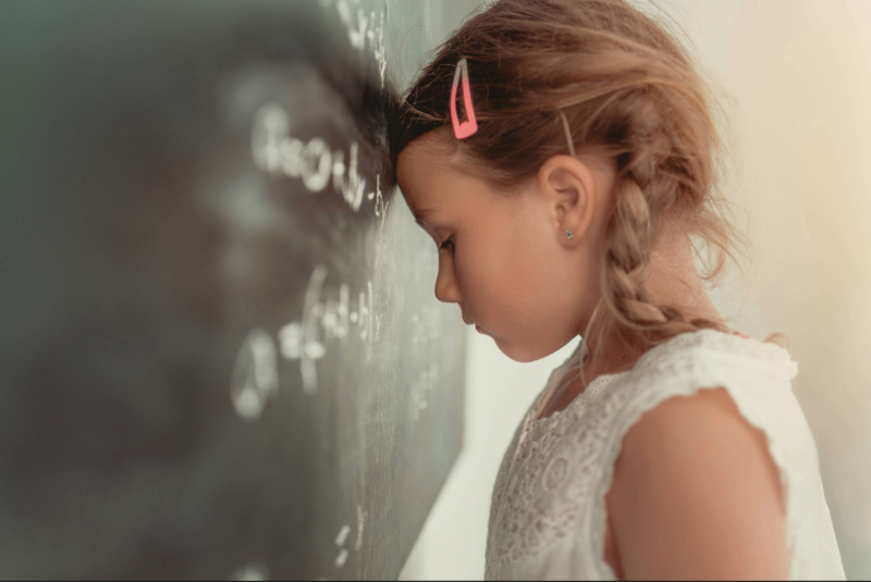 Mädchen lehnt im Klassenzimmer den Kopf an die Tafel, ein Bild für Überforderung, Reizstress und Konzentrationsprobleme bei ADHS in der Schule